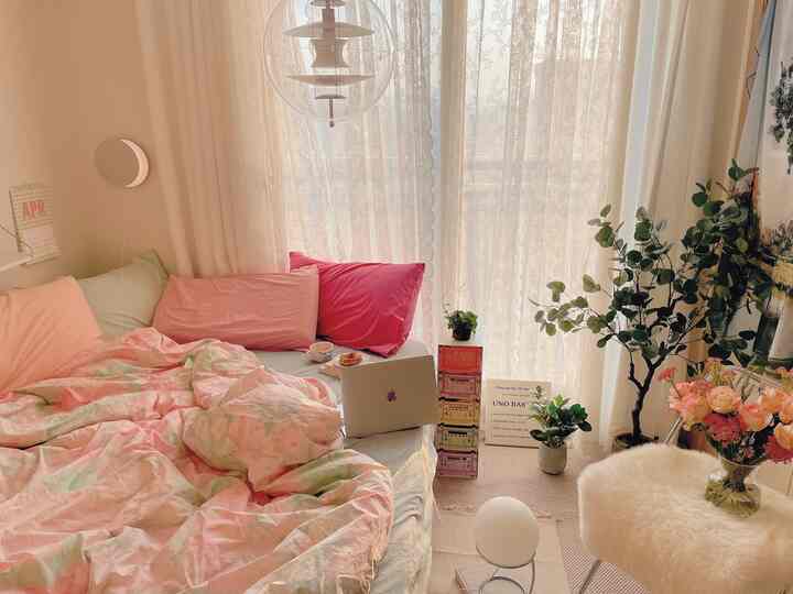 Soft beige and pastel-toned bedroom featuring lace curtains on windows, vases, plants, and pink pillows on the bed creating a cozy atmosphere