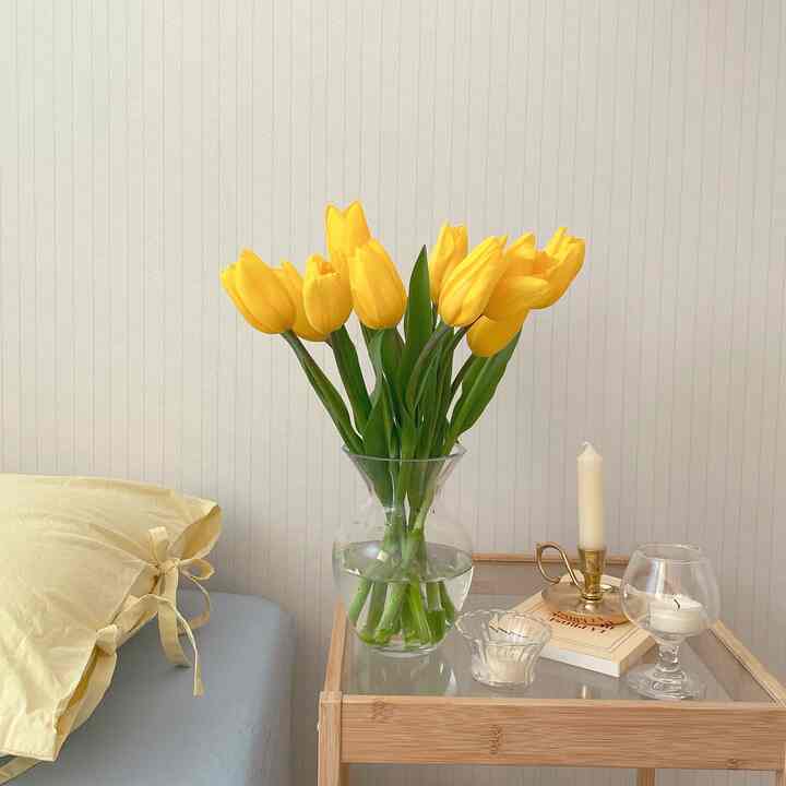 A natural-toned bedroom featuring a nightstand with a vase of yellow tulips and candles, complemented by yellow bedding for a cozy atmosphere