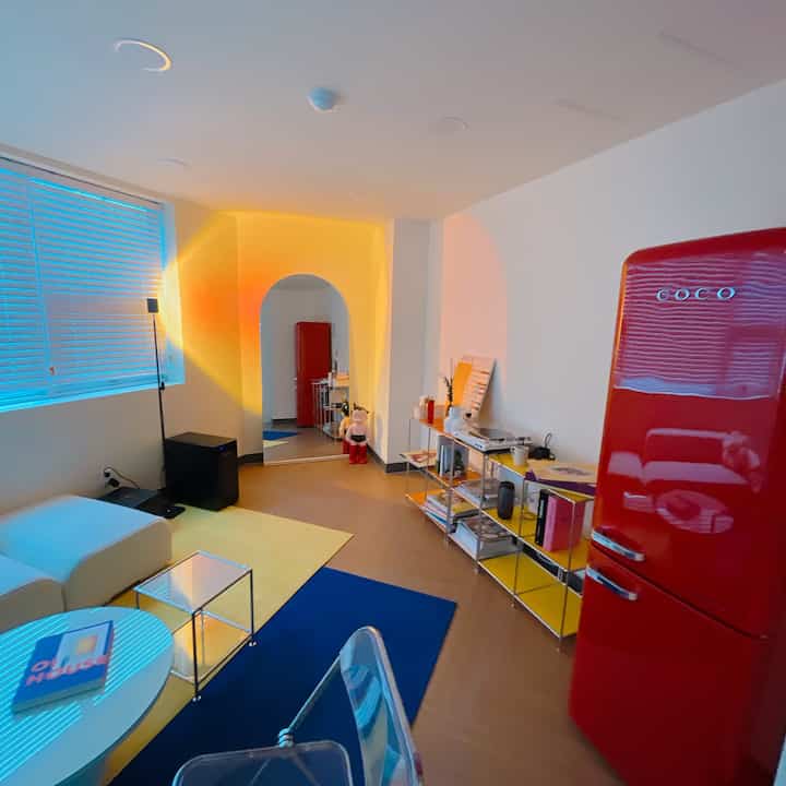 A studio apartment living room with red and blue accents, featuring a white sofa, transparent chair, and retro-style refrigerator in a modern setting
