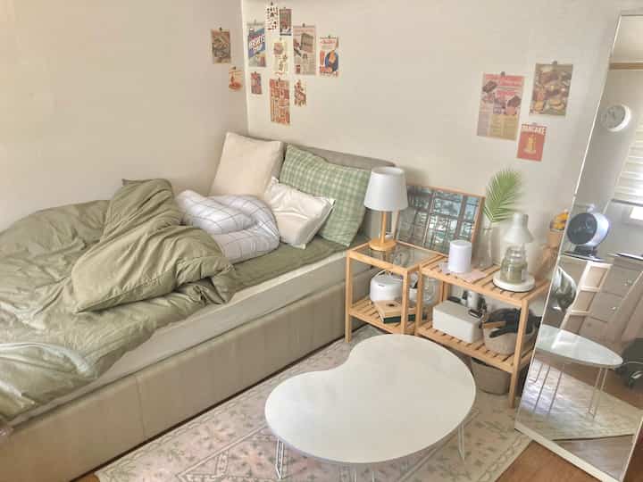 A bedroom space with green and beige tones, featuring natural wooden side tables, cushions, and posters on the wall creating a cozy atmosphere