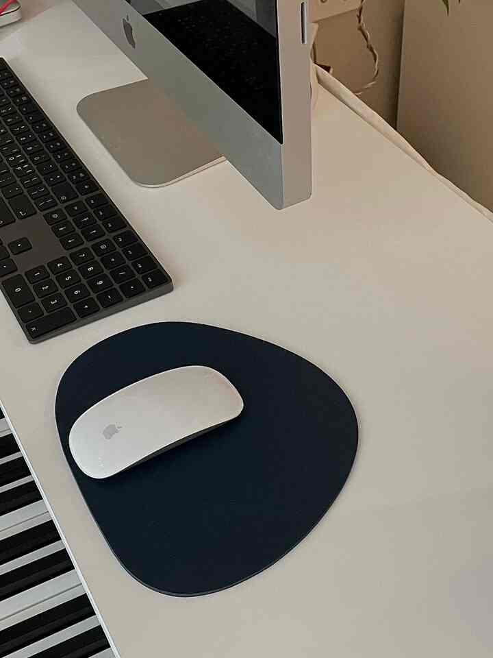 A white and black toned study featuring a long desk with an iMac, keyboard instrument, and navy mouse pad in a neat workspace
