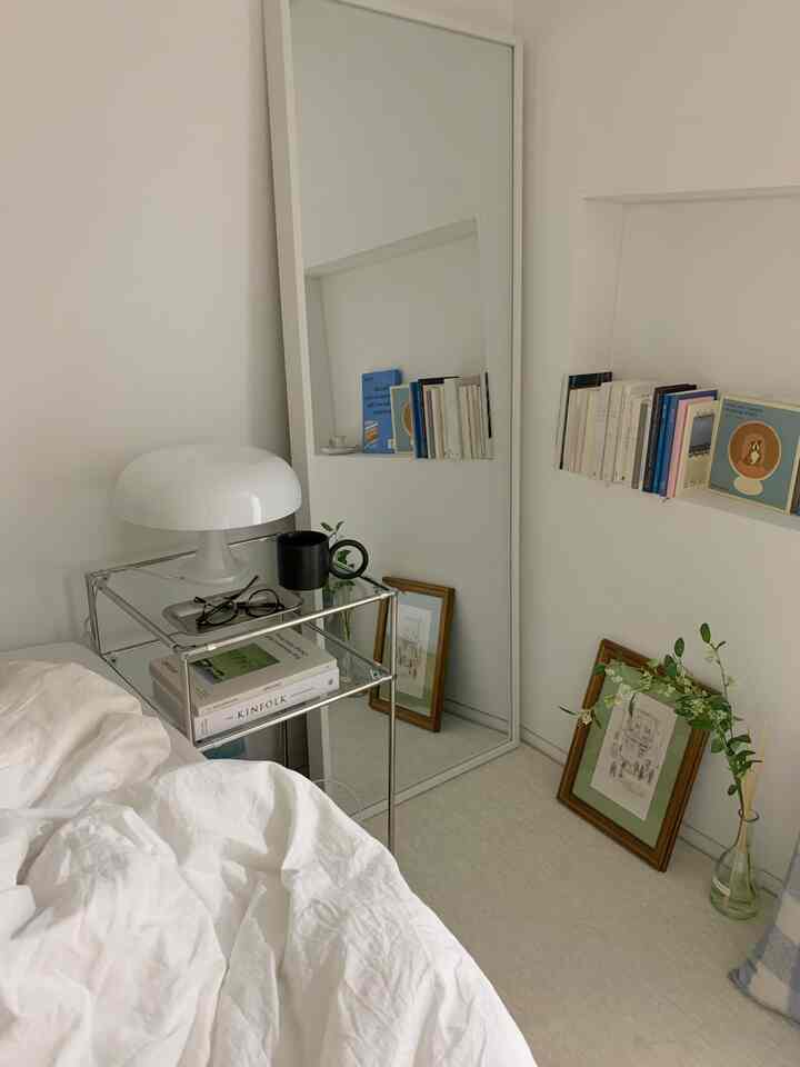 ホワイトのナチュラルトーン寝室、ベッドや全身鏡、本棚、テーブルランプが配置されたシンプルな空間