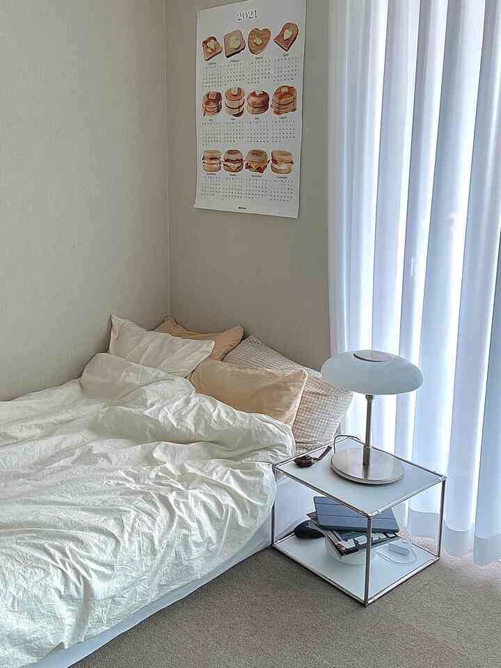 White and beige toned bedroom featuring a poster, curtains, and a table lamp on the nightstand creating a neat and cozy atmosphere