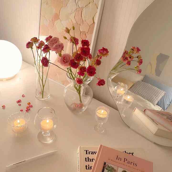 Pastel pink and beige vanity space featuring vases with flowers, a wavy mirror, and lit candles creating a warm cozy atmosphere