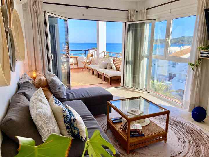 Bright natural-toned living room featuring a gray sectional sofa, rattan coffee table, and large balcony windows with sea view