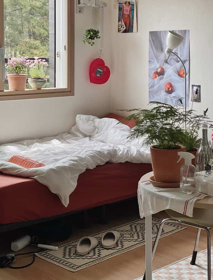 白い寝具と赤いマットレスがあるワンルームの寝室、自然素材と植物が調和した落ち着いた空間