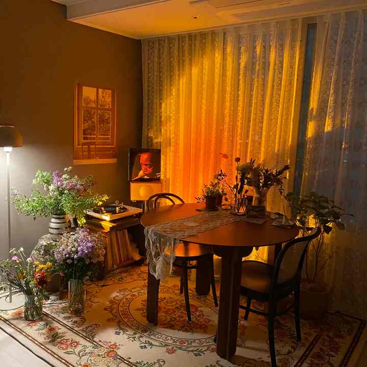 温かみのある黄金色の照明が灯るヴィンテージスタイルのリビング、円形の木製テーブルとペルシャ絨毯、多様な花と植物が調和した雰囲気のあるインテリア