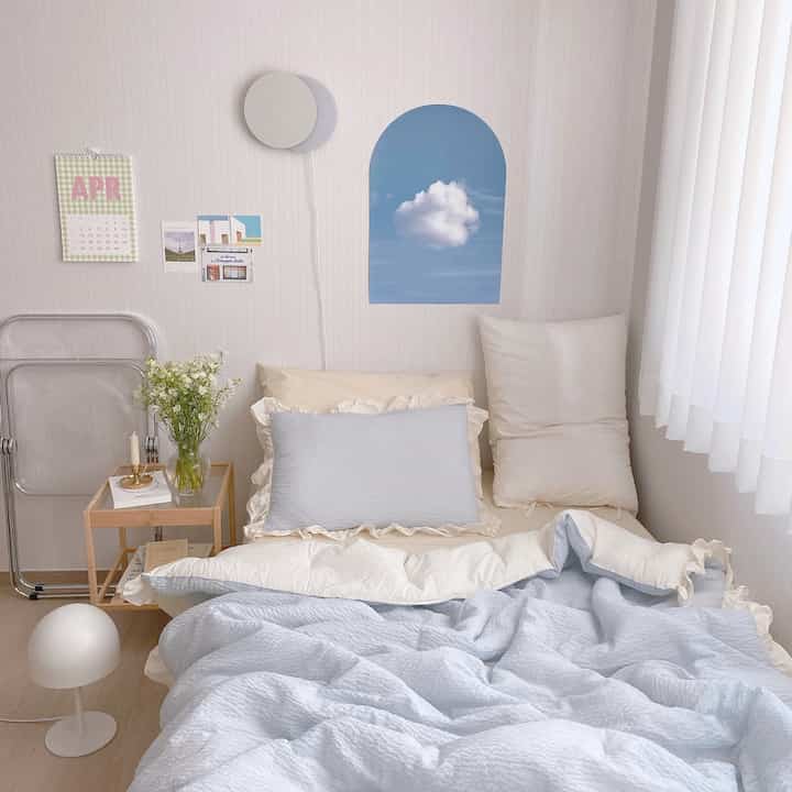 Cozy bedroom with white walls and curtains featuring blue-toned bedding and natural wood nightstand in a natural modern style