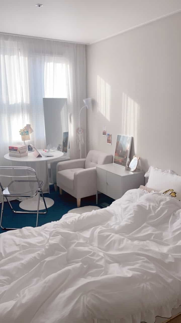 Bright and cozy studio apartment with white bedding, armchair, and round table in natural white and beige tones