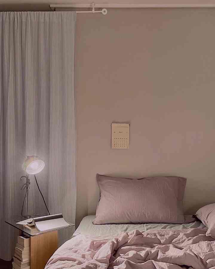 柔らかなベージュ調のワンルーム寝室、ピンクの寝具とガラスのナイトテーブル、テーブルランプが配置されたミニマルなインテリア