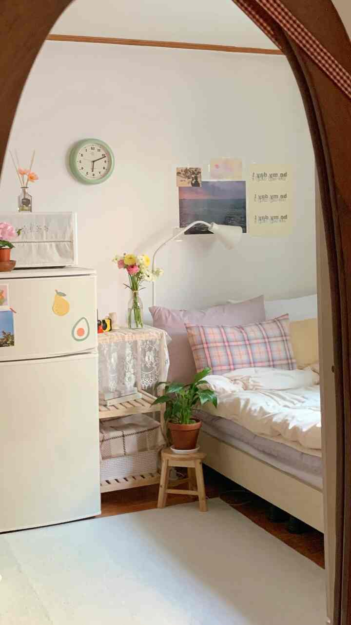 白い壁と木製家具が配置された6坪のワンルーム寝室、ビンテージレースのテーブルクロスとチェック柄のクッションがアクセントの落ち着いた空間