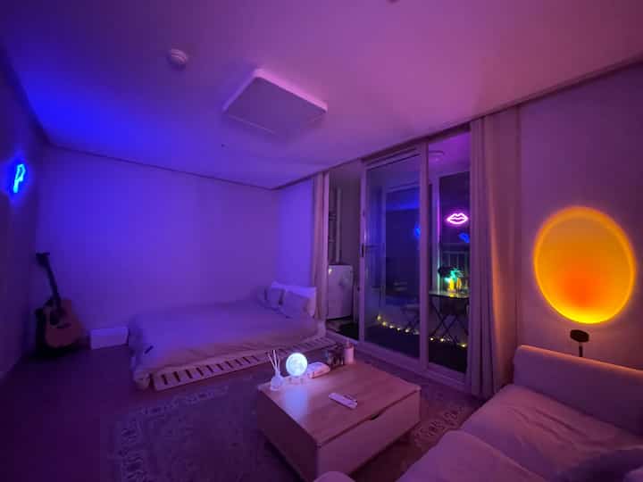 Purple and white toned bedroom-living space featuring wood bed and white sofa, highlighted by neon signs and sunset lighting for a trendy vibe