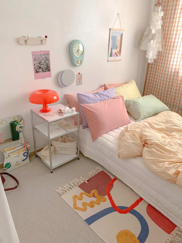 Pastel-toned studio bedroom featuring a white bed with multi-colored pillows and an orange table lamp, creating a cozy atmosphere