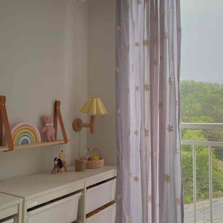 白とナチュラルカラーの子供部屋、チェック柄の花の刺繍カーテンと棚、おもちゃ収納が配置された雰囲気のある空間