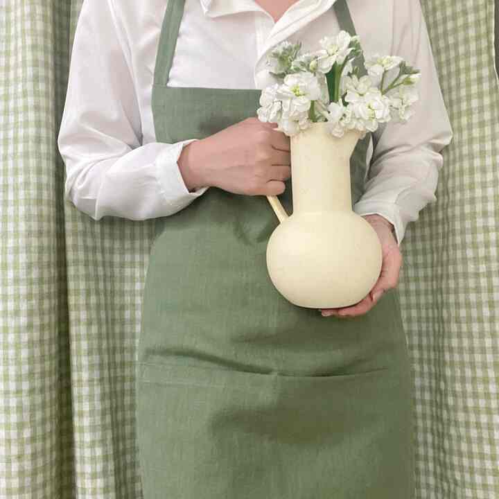 落ち着いた緑のチェックカーテンとグリーンのエプロンを着た人物がベージュのヴィンテージのジャグ花瓶を持つキッチンの雰囲気のあるインテリア