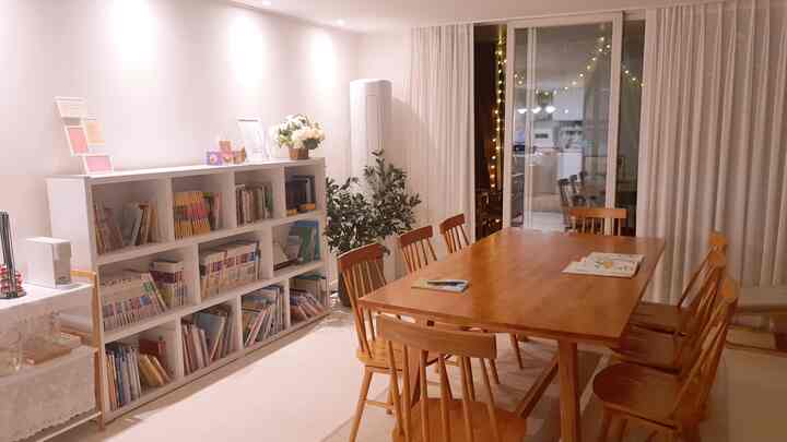 ナチュラルな木目調のリビング兼書斎空間、大きなダイニングテーブルと8脚の椅子、白い本棚が配置された落ち着いたインテリア