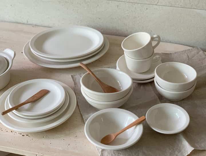 ナチュラルなベージュのテーブルに、ホワイトのセラミック食器セットと木製スプーンが配置された清潔感のある温かみのあるキッチンインテリア