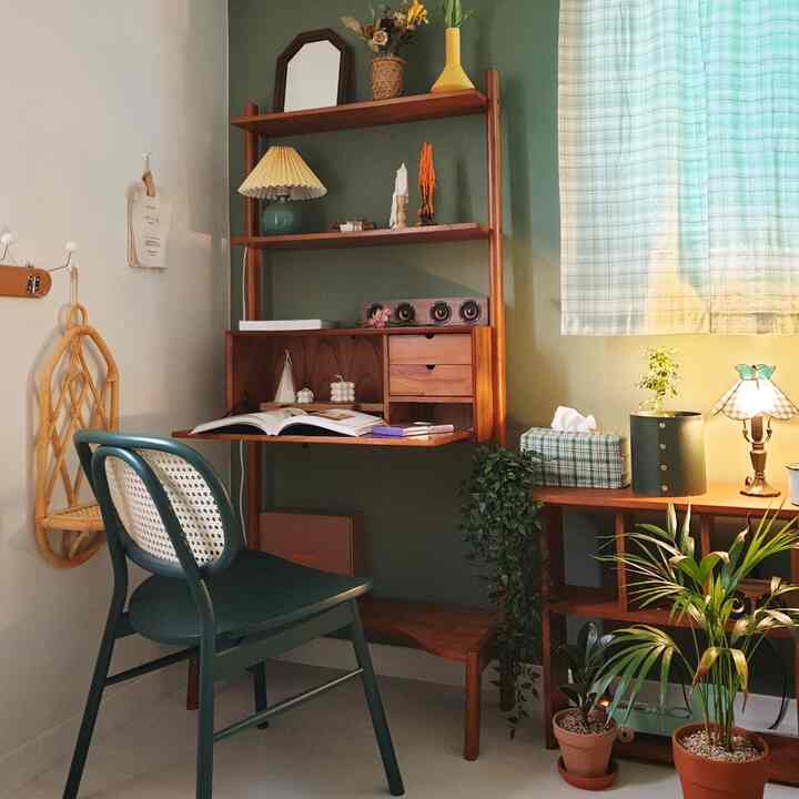 Green-toned vintage mid-century modern home office space featuring wooden desk and chair, assorted plants, and patterned curtain creating a cozy atmosphere