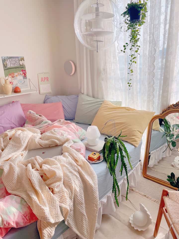 Pastel tones and white bedroom featuring a large bed with multiple pillows, natural light, and greenery, creating a cozy atmosphere