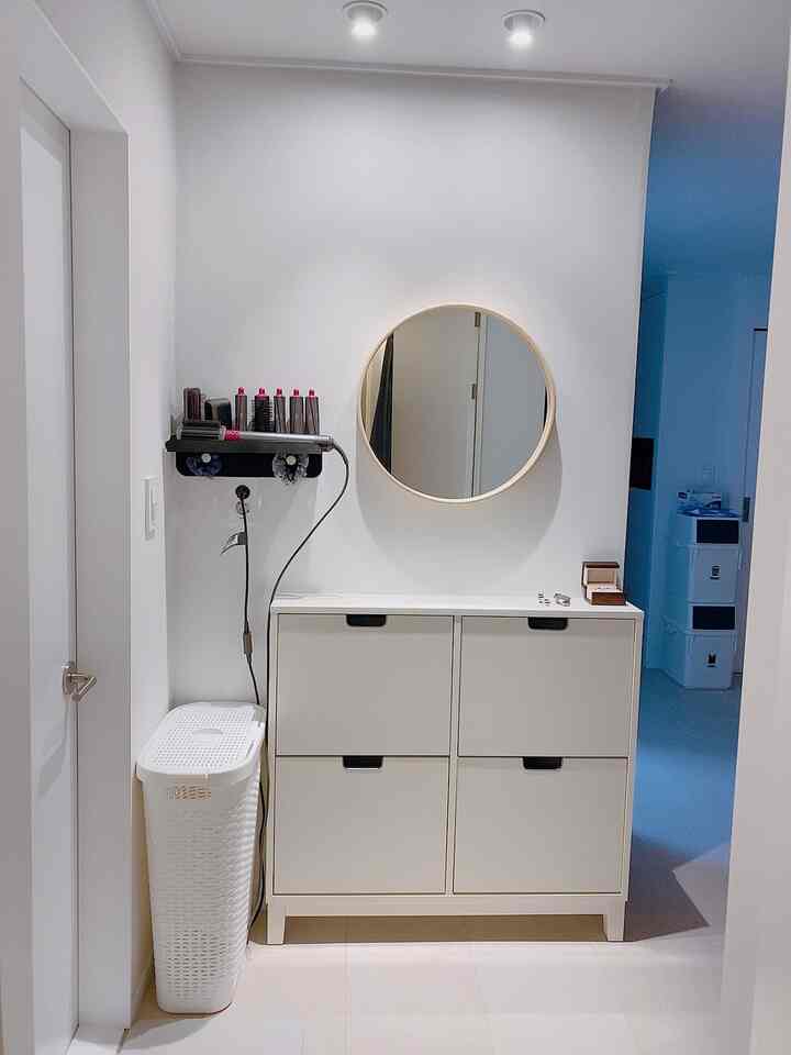 White-toned narrow space featuring a slim cabinet dresser, wood round mirror, and white laundry basket, creating a practical vanity area