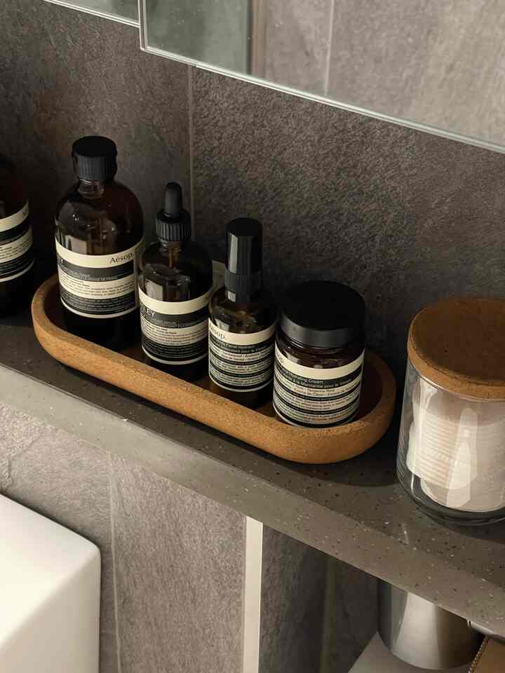 ナチュラルなブラウンとグレーの色調のバスルーム、鏡の下の棚に整然と並べられたイソップ製品が特徴的な清潔感のあるインテリア