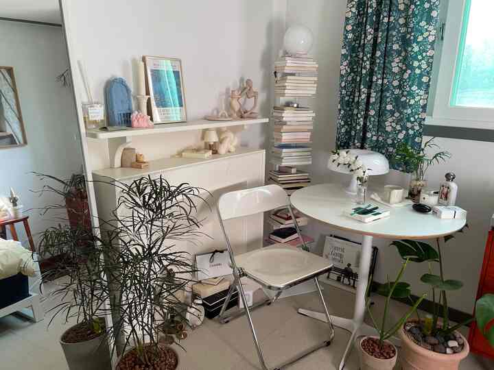 白と緑のトーンの狭い部屋、丸テーブルと折りたたみ椅子、植物と積まれた本が配置された雰囲気のあるインテリア