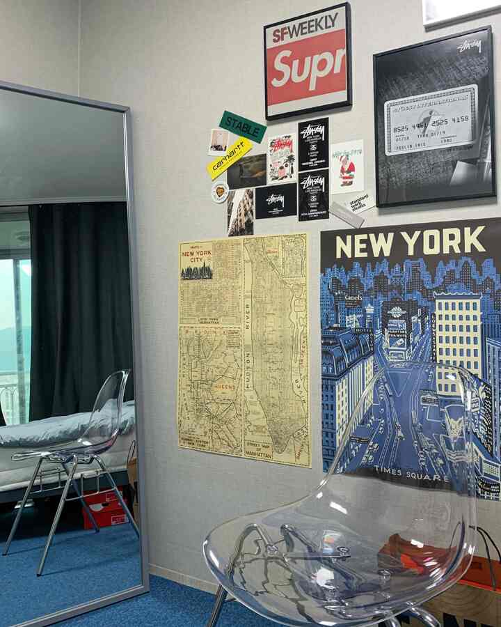 Compact room with blue carpet and beige wallpaper featuring a mirror and transparent chair against a wall decorated with New York posters