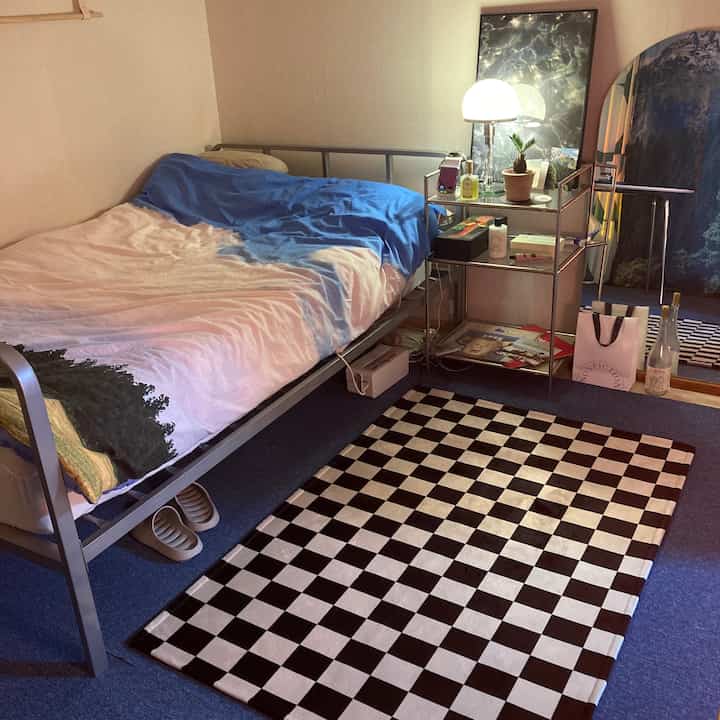 A compact bedroom with beige walls and blue carpet, featuring a single bed, checkered rug, side table with table lamp creating a cozy atmosphere