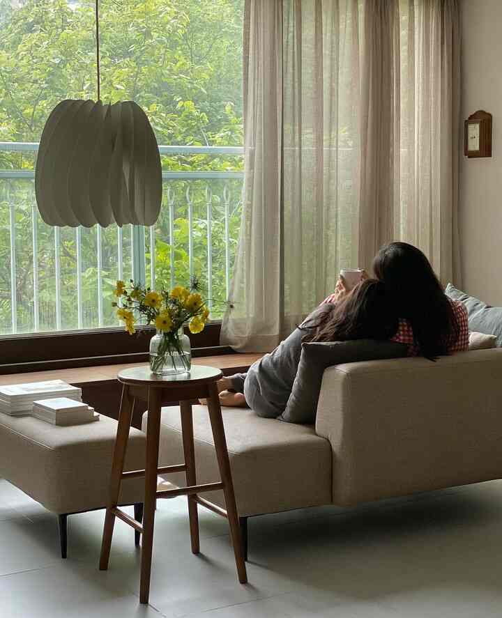 A cozy natural living room with beige and wood tones featuring a two-seater sofa and wooden stool near a large window with light curtains