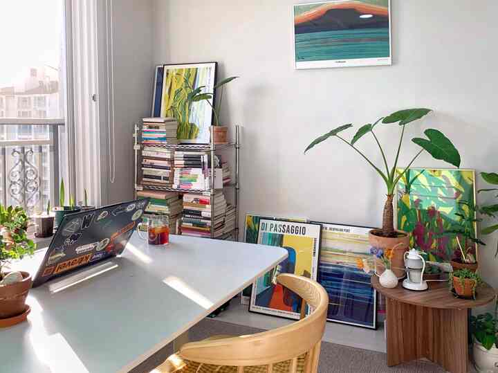 A white and wood tone small home study featuring a desk, chair, numerous plants, and framed posters, creating a cozy home office atmosphere