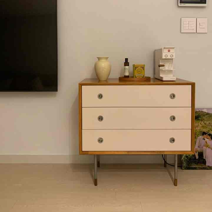 White-walled living room with a wood-toned framed dresser placed in a compact space, topped with a coffee machine and vase, creating a neat home cafe area