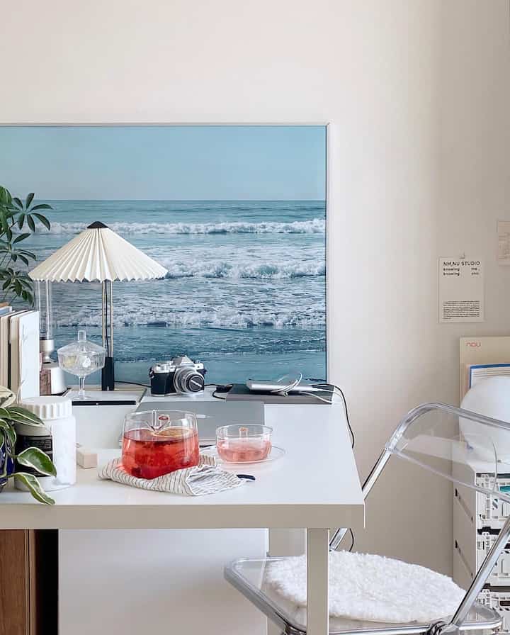 白と透明素材を基調としたホームオフィス空間で、海のポスターと植物が配され落ち着いた清潔感のある作業環境