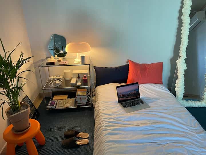 ホワイトとオレンジのトーンが映えるワンルーム寝室、シンプルな寝具とオレンジのスツール、植物、フロアランプが配置されたミニマルな空間