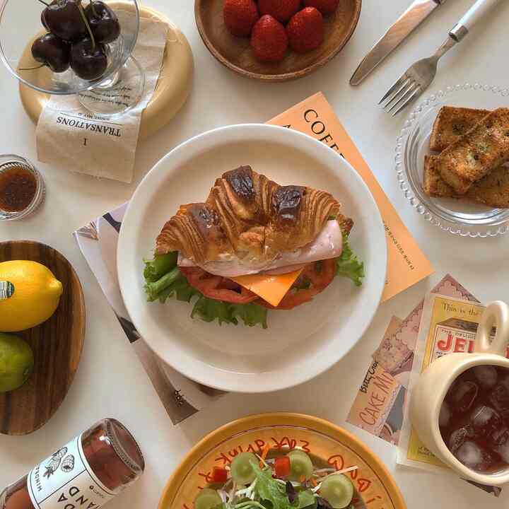 A white-toned kitchen table featuring a croissant sandwich, fruit bowls, and iced tea in a home cafe setting