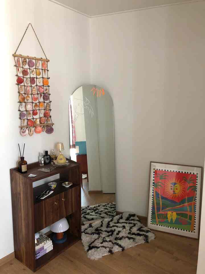 ブラウンの木目調家具と白い壁のワンルーム、全身鏡とラグ、カラフルな額縁が配置されたナチュラルモダンな雰囲気のインテリア