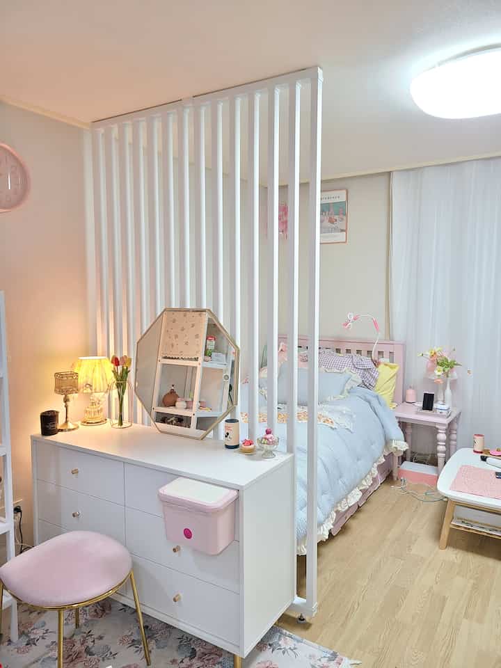 Pastel-toned studio bedroom with pink furniture, featuring a white room divider creating a cozy separated space