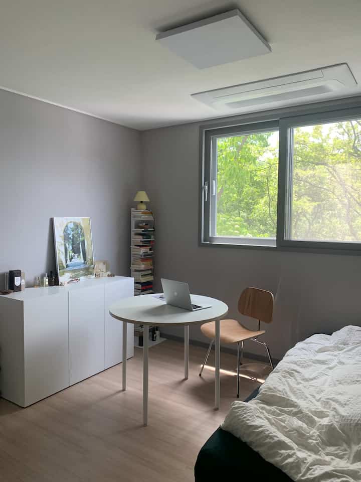 A white and wood tone modern single household bedroom with remote work setup, featuring a round table, bookshelf, and large window