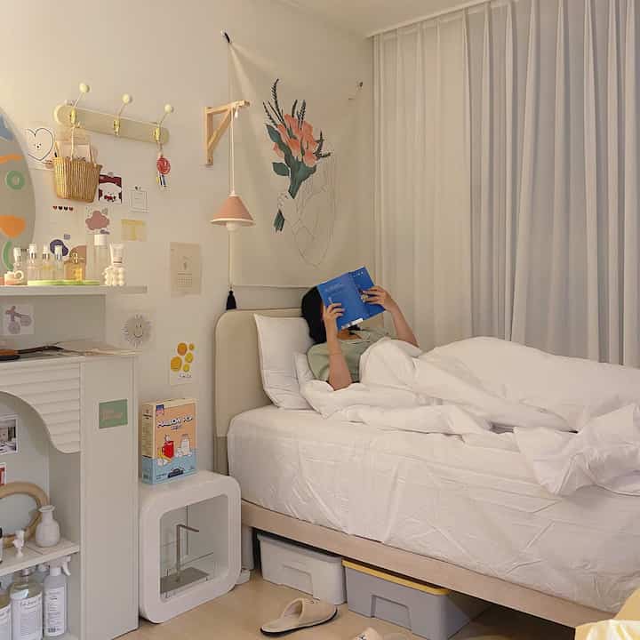 A cozy small bedroom with white and natural wood tones, featuring a poster, pendant light, and a person reading comfortably in bed