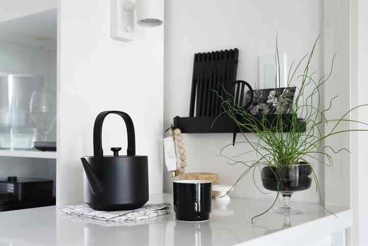 Modern kitchen with a white backdrop and black accents featuring a black oriental teapot and coffee cup centered on the counter, with a green plant to the right creating a clean home cafe space.