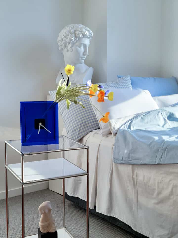 Sky blue and white toned bedroom featuring light blue bedding, white plaster bust, and a transparent blue clock on a modern side table