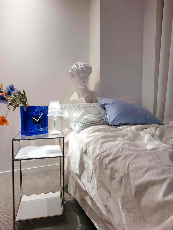 A white and blue toned bedroom featuring a nightstand, table lamp, and white objet with a simple atmosphere