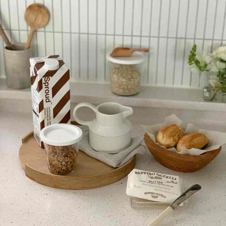 ホワイトとナチュラルブラウンの色調のキッチン、木製トレイ、パン、ガラス瓶、バターが整然と配置された雰囲気のあるおうちカフェのインテリア