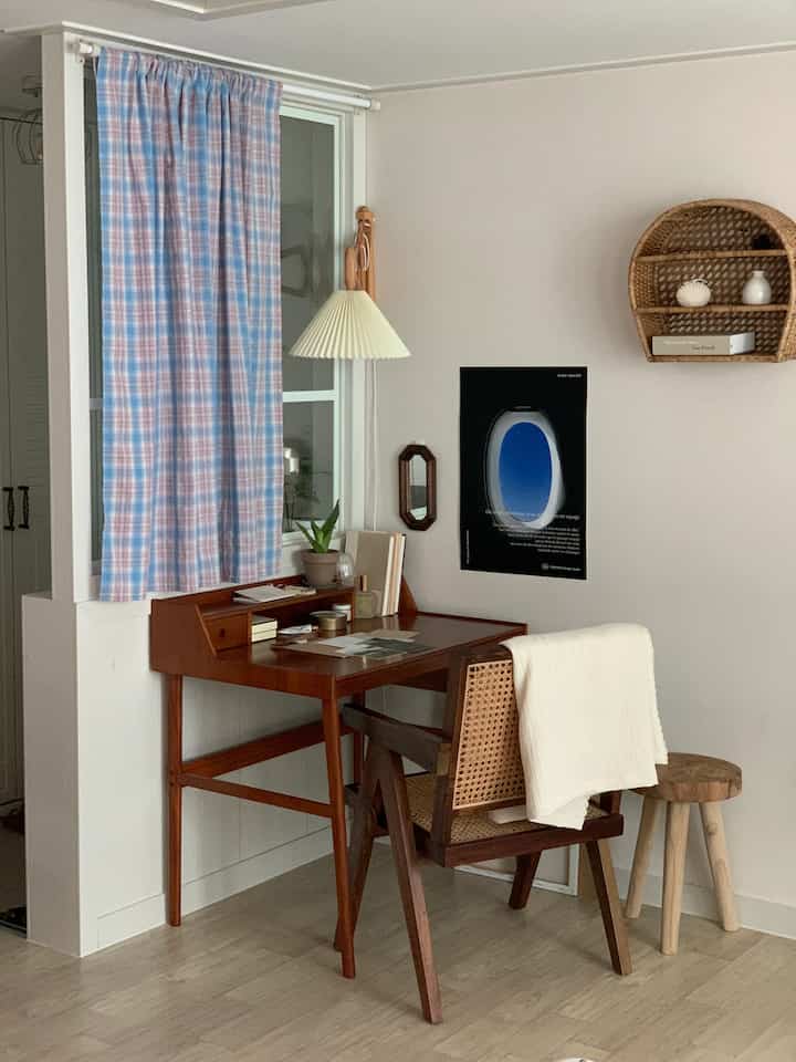 Natural tone living room with white walls, brown wooden desk, rattan shelf, and blue-pink plaid curtain in home office corner