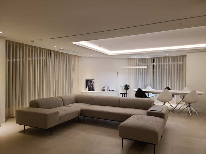Living and dining room in soft beige and white tones, featuring a large modern sectional sofa and an elongated dining table with a minimalist design