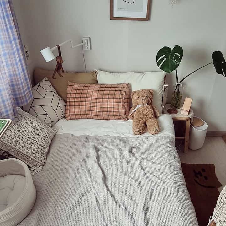 ベージュとブラウンのトーンの寝室、ベッドにテディベアとクッション、左側に紫のチェック柄カーテンがある自然な雰囲気のインテリア