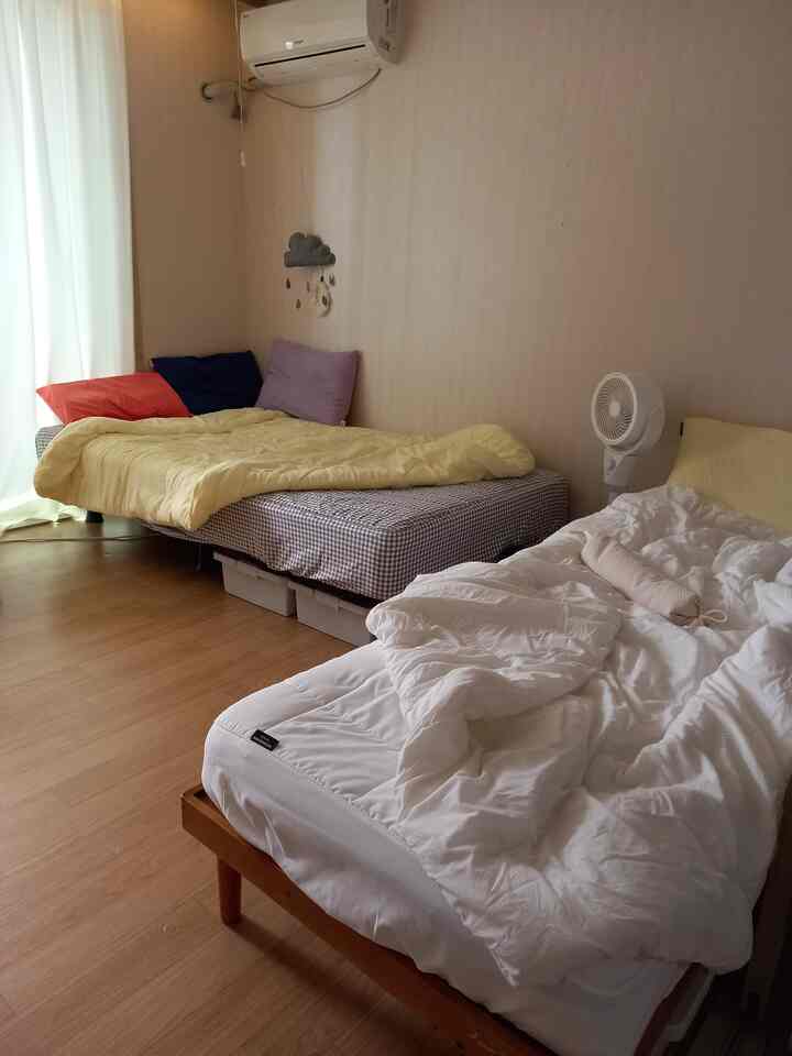 A compact bedroom with beige walls and hardwood flooring featuring two beds arranged in an L-shape and colorful pillows creating a cozy atmosphere