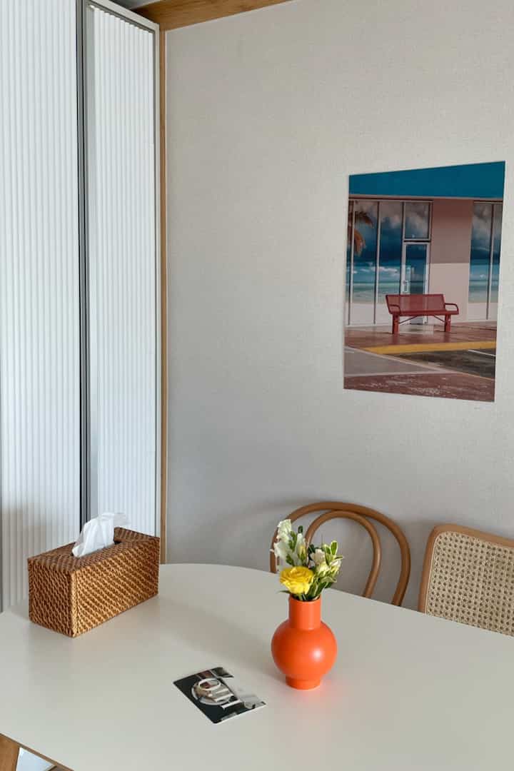 Natural dining room with white walls and wood-tone chairs, featuring an orange vase and framed picture as charming accents