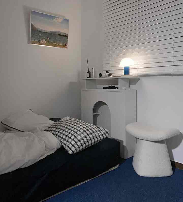Blue and white toned bedroom featuring single bed, textured stool, blinds, and framed artwork in a modern cozy setting
