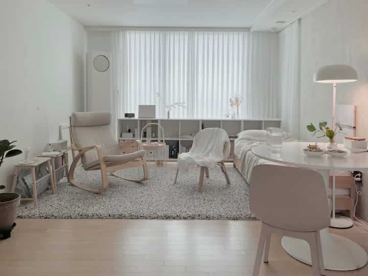 Light beige and white toned studio apartment living room featuring a daybed, armchair, and round dining table with a calm atmosphere