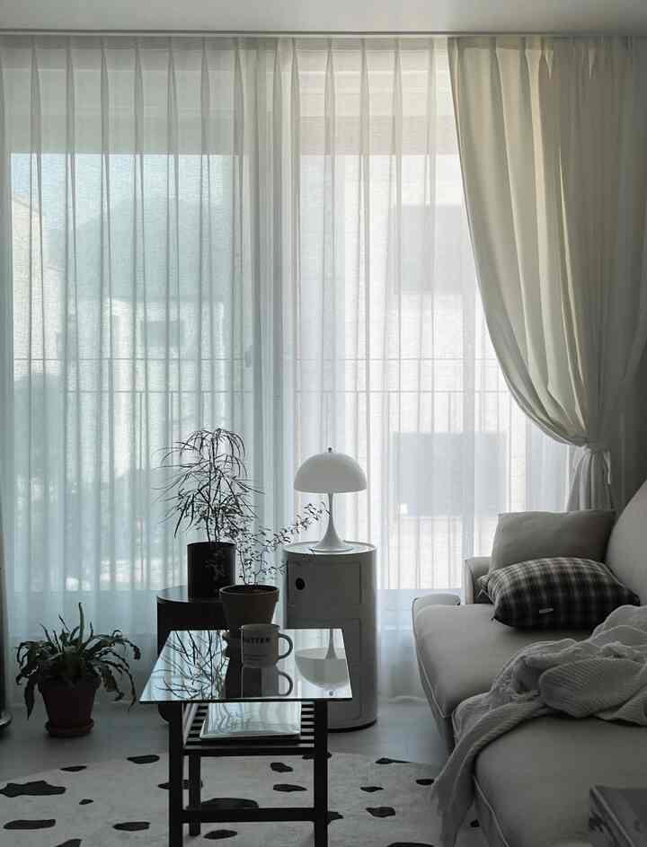 ホワイトとベージュの色調のリビング、ガラスのコーヒーテーブルと円形ラグ、植物とシンプルな家具が配置された落ち着いた空間